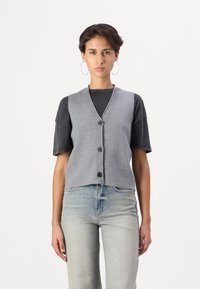 Gilet grigio lavorato a maglia con scollatura a V, cinque bottoni frontali e orlo corto. Indossato sopra una maglietta nera e jeans chiari.