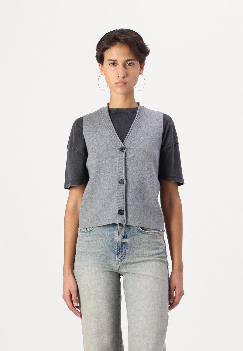 Gilet grigio lavorato a maglia con scollatura a V, cinque bottoni frontali e orlo corto. Indossato sopra una maglietta nera e jeans chiari.
