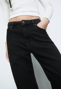 Pantaloni in denim neri con taglio dritto, dettagli con borchie e una vita classica con chiusura a bottone. Tessuto testurizzato con motivo a punti.