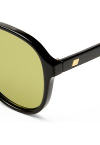 Κοντινό πλάνο μαύρων γυαλιών ηλίου με φακούς με κίτρινο-πράσινη απόχρωση και μικρή χρυσή λεπτομέρεια διαμαντιού στο μπράτσο του σκελετού, σε λευκό φόντο.