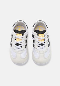 Chaussures de sport blanches et noires en matériaux synthétiques et daim, munies de trois bandes emblématiques, d'un bout rond et d'accents noirs texturés.