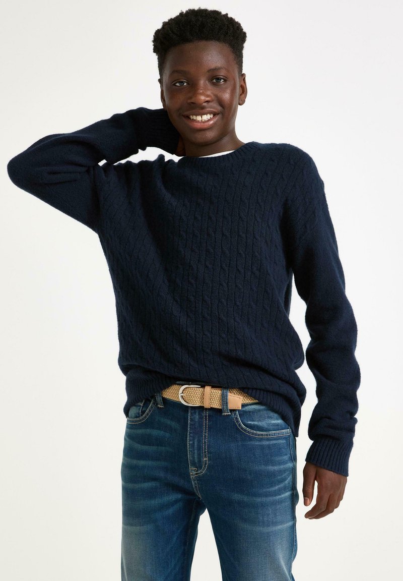 Granatowy sweter w warkoczowy wzór z ribbowymi mankietami i dołem, w połączeniu z niebieskimi dżinsami i plecionym brązowym paskiem. Prosty i teksturowany design.