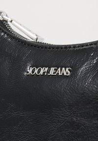 Mustaherkkä nahkainen pussi, jossa on hopeinen vetoketju ja metallinen logo, jossa lukee "JOOP! JEANS" edessä.