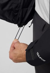 Veste imperméable noire avec cordons de serrage réglables à l'ourlet. Présente une manche texturée avec le logo "TEXAPORE". Main tenant le cordon.
