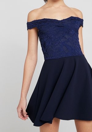 Navy blue off-shoulder dress with fitted floral lace bodice and flared skirt worn by a standing person against a plain background.