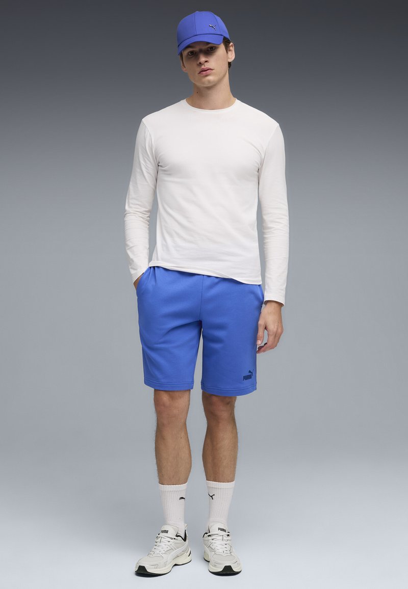 Jeune homme portant une casquette bleue, une chemise blanche à manches longues, un short bleu, des chaussettes blanches et des baskets blanches, debout devant un fond gris.