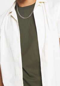 Weißes kurzärmliges Button-Up-Hemd über einem olivgrünen T-Shirt, ergänzt durch eine silberne Kettenhalskette. Das Hemd hat eine weiche Textur und eine lockere Passform.