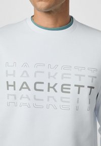 Uomo che indossa un felpa bianca con la scritta ripetuta "HACKETT" in diverse tonalità e stili sul petto.
