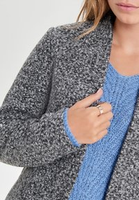 Kvinna som bär en texturerad grå kappa över en blå stickad tröja, som håller i kappans krage med vänster hand och visar en enkel metallring.