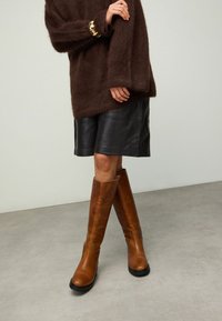 Bruine pluizige trui, zwarte leren rok en hoge bruine leren laarzen met een zwarte zool. Gezellige, gestructureerde materialen met gestroomlijnde vormen.