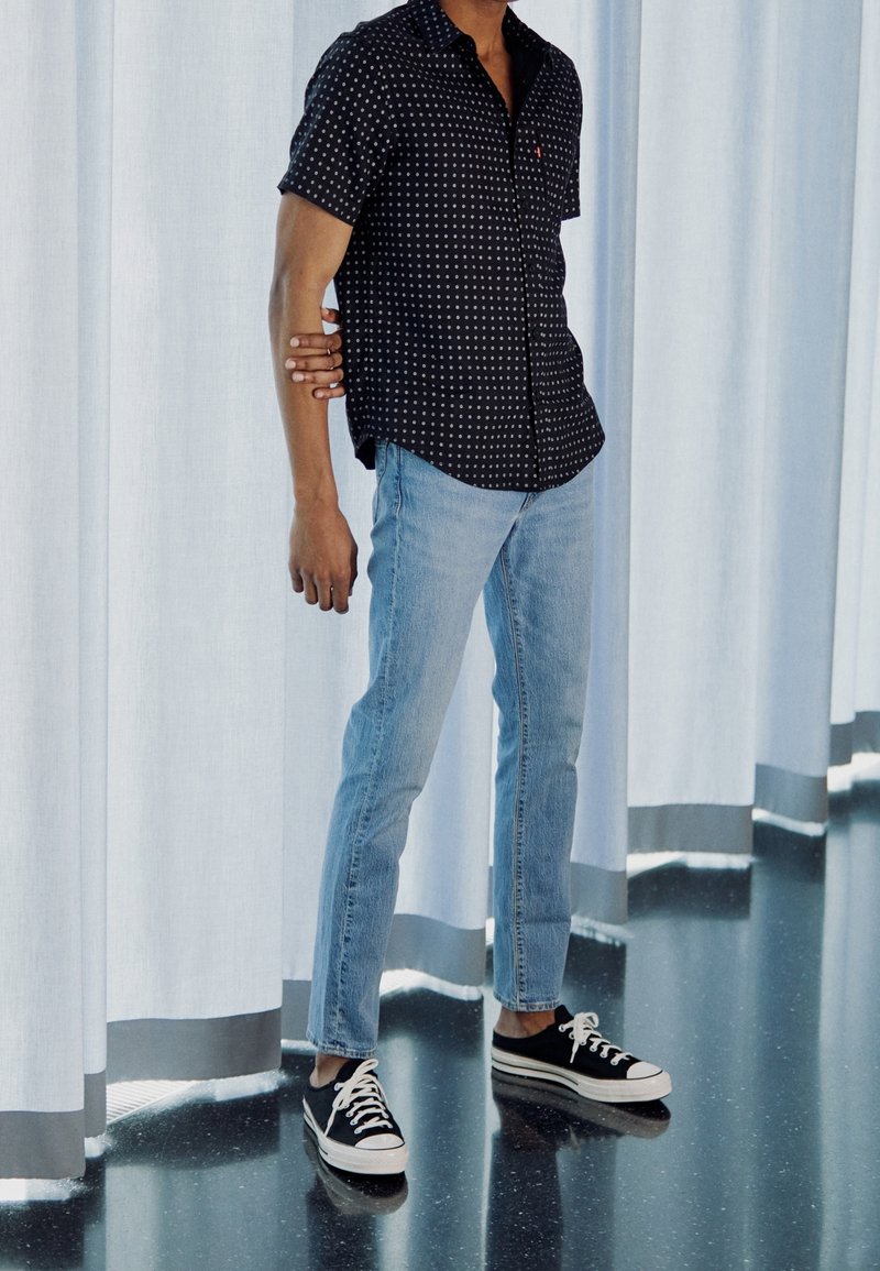 Personne portant une chemise noire à manches courtes à motifs, un jean bleu clair et des baskets noires, debout avec le bras plié derrière le dos devant des rideaux blancs.