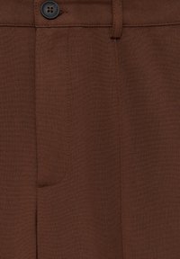 Detailní záb�ěr hnědých tkaninových kalhot ukazující pás, knoflík, poutko na opasek a prošité detaily kapsy.