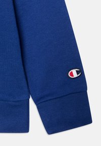 Puño de sudadera azul con textura de canalé, que presenta un logotipo bordado con acentos rojos y blancos sobre un óvalo negro.