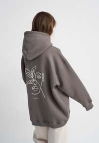 Felpa grigia oversize con zip frontale, caratterizzata da un disegno a linee bianche di un volto e foglie sul retro. Tessuto morbido, design casual.