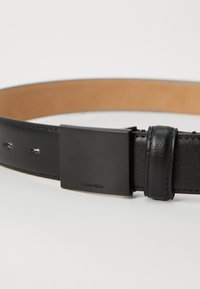 Cinturón de cuero negro con un acabado liso, que presenta una hebilla cuadrada negra grabada con "Calvin Klein" y costuras visibles a lo largo de los bordes.