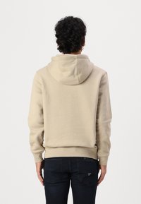 Beige hoodie with a kangaroo pocket and adjustable drawstring hood, made from soft material, paired with dark denim jeans.