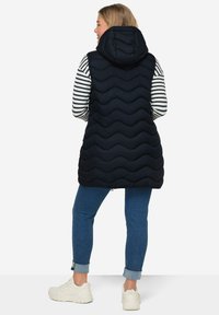 Marinblå pufferväst med huva, med ett vågmönster, dragkedjestängning och sidofickor, bärs över en randig långärmad skjorta och jeans.