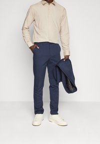 Beige Hemd mit langen Ärmeln, blaue Hose und weiße Sneaker. Der marineblaue Blazer wird in einer Hand gehalten, mit einem dunklen Innenfutter.