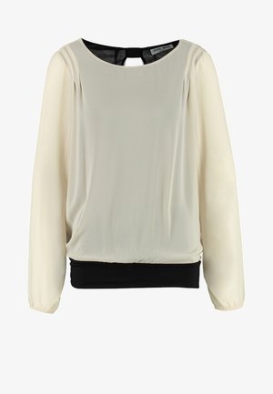 Vrouw met een zwarte leren jas, beige broek, witte blouse en zwarte enkellaarsjes, staand met één arm lichtjes uitgestrekt.