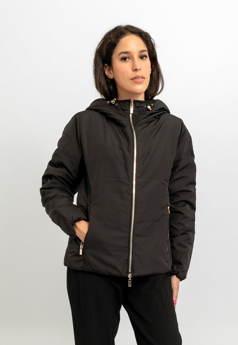 Giacca a vento nera imbottita con cappuccio, cerniera frontale e dettagli in metallo dorato. Presenta polsini elastici e un materiale liscio e resistente.