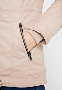 Jasnoróżowa kurtka z zamykaną kieszenią na suwak, ściągaczami przy rękawach i gładką fakturą. Widoczne szwy oraz subtelny, elastyczny pas w talii.