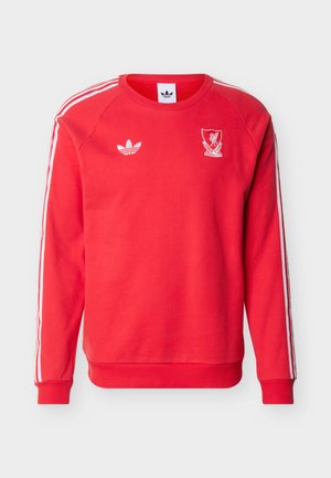 Crvena dugih rukava Adidas majica s bijelim prugama na rukavima, Adidas logotipom na lijevoj strani prsa i grbom Liverpool nogometnog kluba na desnoj strani prsa.