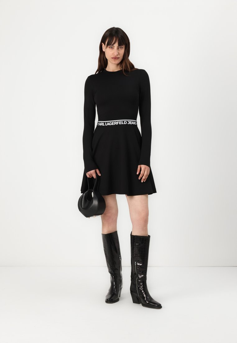 Robe noire à manches longues avec jupe évasée et une ceinture de marque blanche. Associée à des bottes noires en cuir verni à talons et un petit sac à main noir.