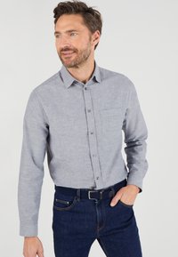 Camicia button-up grigia con tessuto testurizzato, maniche lunghe e taschino sul petto, abbinata a jeans in denim blu scuro e una cintura.
