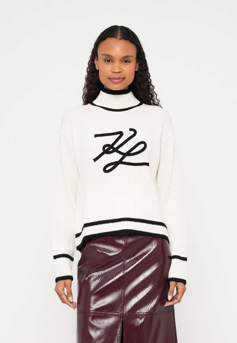 Bela rebrasta majica s črnimi poudarki in izrazitim črnim logotipom na sprednji strani. Model nosi sijočo bordo krilo, ki doda kontrast.