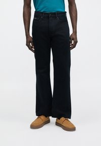 Čierne džínsy s širokými nohavicami a vysokým pásom, so zdôrazneným kontrastným prešívaním a prednými vreckami, skombinované s hnedými semišovými topánkami a tyrkysovou košeľou.