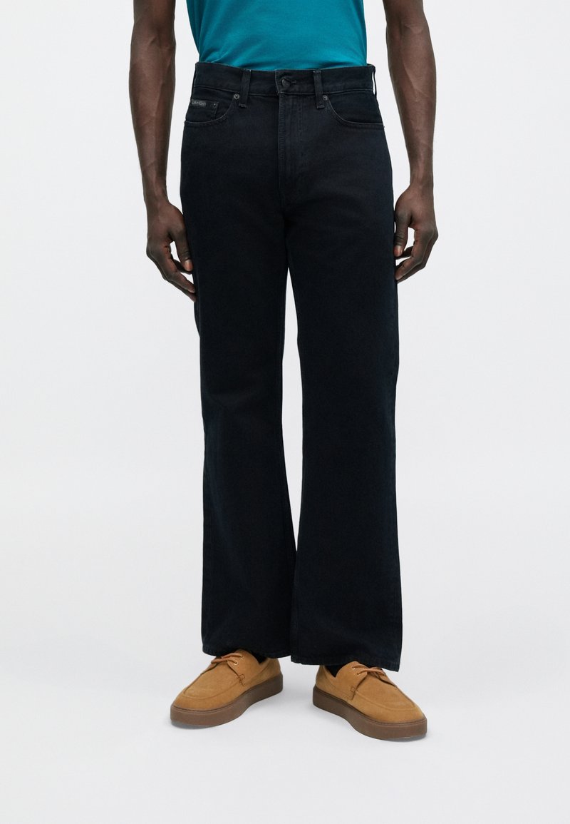 Čierne džínsy s širokými nohavicami a vysokým pásom, so zdôrazneným kontrastným prešívaním a prednými vreckami, skombinované s hnedými semišovými topánkami a tyrkysovou košeľou.