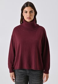 Burgunderfarbener Rollkragenpullover aus weichem Stoff. Hat einen hohen, gerippten Kragen und eine lockere Passform mit langen Ärmeln. Wird mit dunklen Jeans getragen.