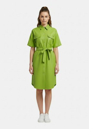 Donna in piedi, indossa un vestito a maniche corte verde acceso con bottoni, tasche e cintura annodata in vita, abbinato a sneakers bianche.