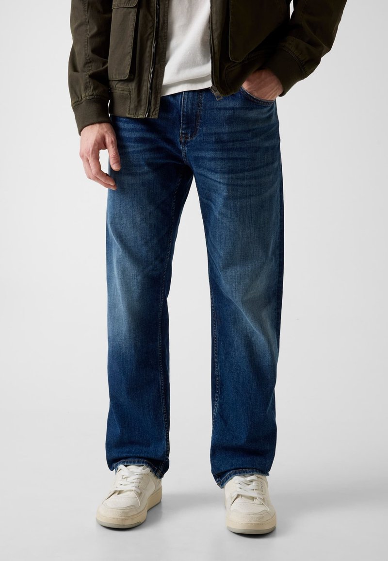 Homme portant un jean bleu droit, des baskets blanches, une chemise blanche et une veste vert foncé avec une main dans la poche.