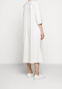 Bílé šaty ke kolenům s třičtvrtinovými rukávy, vyrobené z hladkého materiálu, s jemnými sklady vzadu a bočními rozparky. V kombinaci s bílými teniskami.