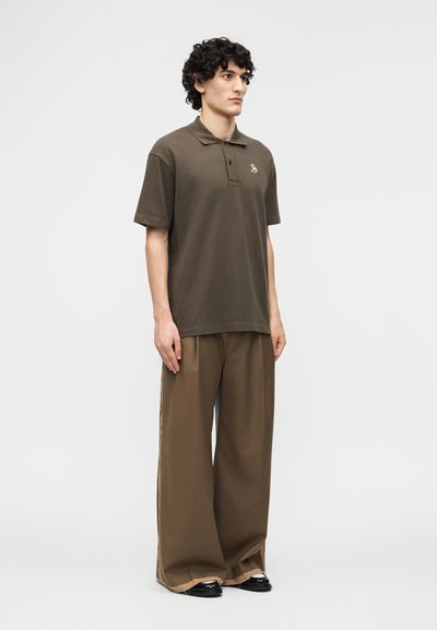 Νεαρός άνδρας με σγουρά μαλλιά φοράει σκούρο ελαιδί πόλο πουκάμισο με μικρό κεντημένο αλεπούδι και καφέ παντελόνι με φαρδιά μπατζάκια μπροστά σε απλό φόντο.