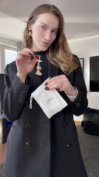 Gullfarget charm-tilbehør med detaljert design, vist med en hvit snorpose merket "Furla." Modellen har på seg en svart frakk.