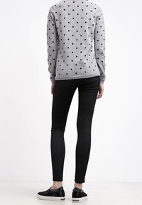 Personne portant une chemise grise à manches longues avec des pois noirs, un pantalon skinny noir et des chaussures noires sans lacets, debout dos à l'objectif.