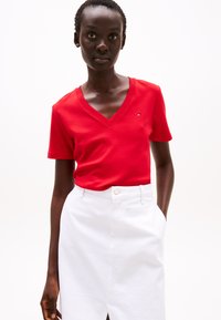 Raudonas V formos kaklo marškinėlis iš minkštos medvilnės, trumpomis rankovėmis, derinamas su baltu džinsiniu sijonu su priekinėmis skeltomis ir šoninėmis kišenėmis.