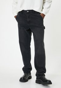 Pantalones cargo de mezclilla negra con un corte recto, bolsillos frontales y detalles de costura visibles. Combinados con botas negras de suela gruesa.