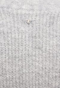 Jasnoszary sweter z dzianiny z teksturowanym wzorem i małą, okrągłą srebrną metką przy dekolcie.