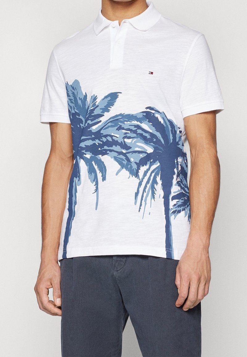 Polo blanc avec un motif de palmier bleu sur le devant. Présente un col et des manches courtes, fabriqué en coton doux.