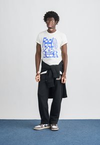 Baltas grafikos marškinėliai su mėlynu dizainu, juodos kelnės su logotipu, juodas džemperis surištas aplink juosmenį, pilki ir balti sportbačiai.