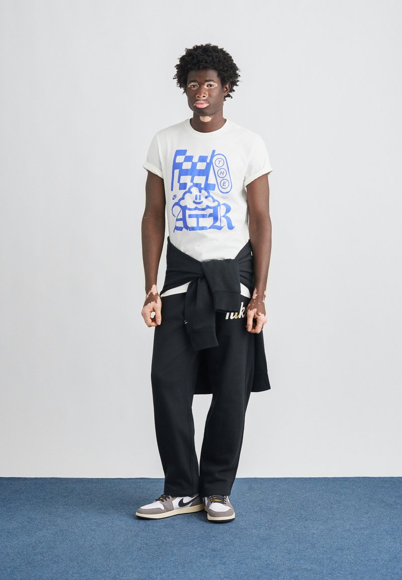 T-shirt graphique blanc avec motif bleu, pantalon de survêtement noir avec logo, sweat-shirt noir noué autour de la taille, baskets gris et blanc.