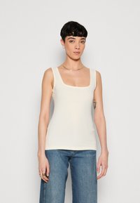 Débardeur crème à col carré, fabriqué en tissu lisse. Il présente des bretelles larges et un design ajusté, associé à un jean bleu.