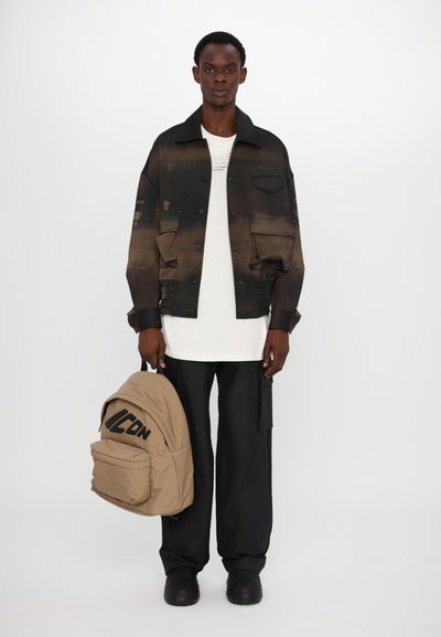 Schwarze und braune Gradient-Jeansjacke mit Fronttaschen, weißem T-Shirt, lockeren schwarzen Hosen und einem sandfarbenen Rucksack mit dunkler Schrift.