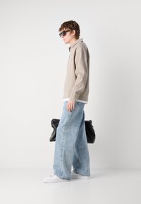Pulover texturat de nuanță bej deschis, blugi de denim lejeri de nuanță albastru deschis, adidași albi și geantă neagră. Modelul poartă ochelari de soare închiși la culoare, văzut din profil.