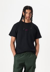Svart bomull t-shirt med en liten lila grafisk logotyp på bröstet, kombinerad med gröna cargobyxor med sidofickor och elastisk midja.