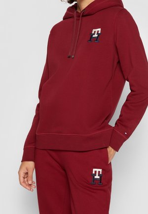 Burgundowy hoodie z kieszenią z przodu, kapturem ze sznurkiem i haftowanym logo w kremowym i granatowym kolorze. Miękka tkanina z prążkowanymi mankietami i dolnym brzegiem.