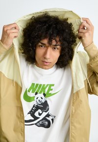 Light beige jacket with hood, white t-shirt featuring green Nike logo and panda graphic, curly dark hair, nose ring, neutral background.